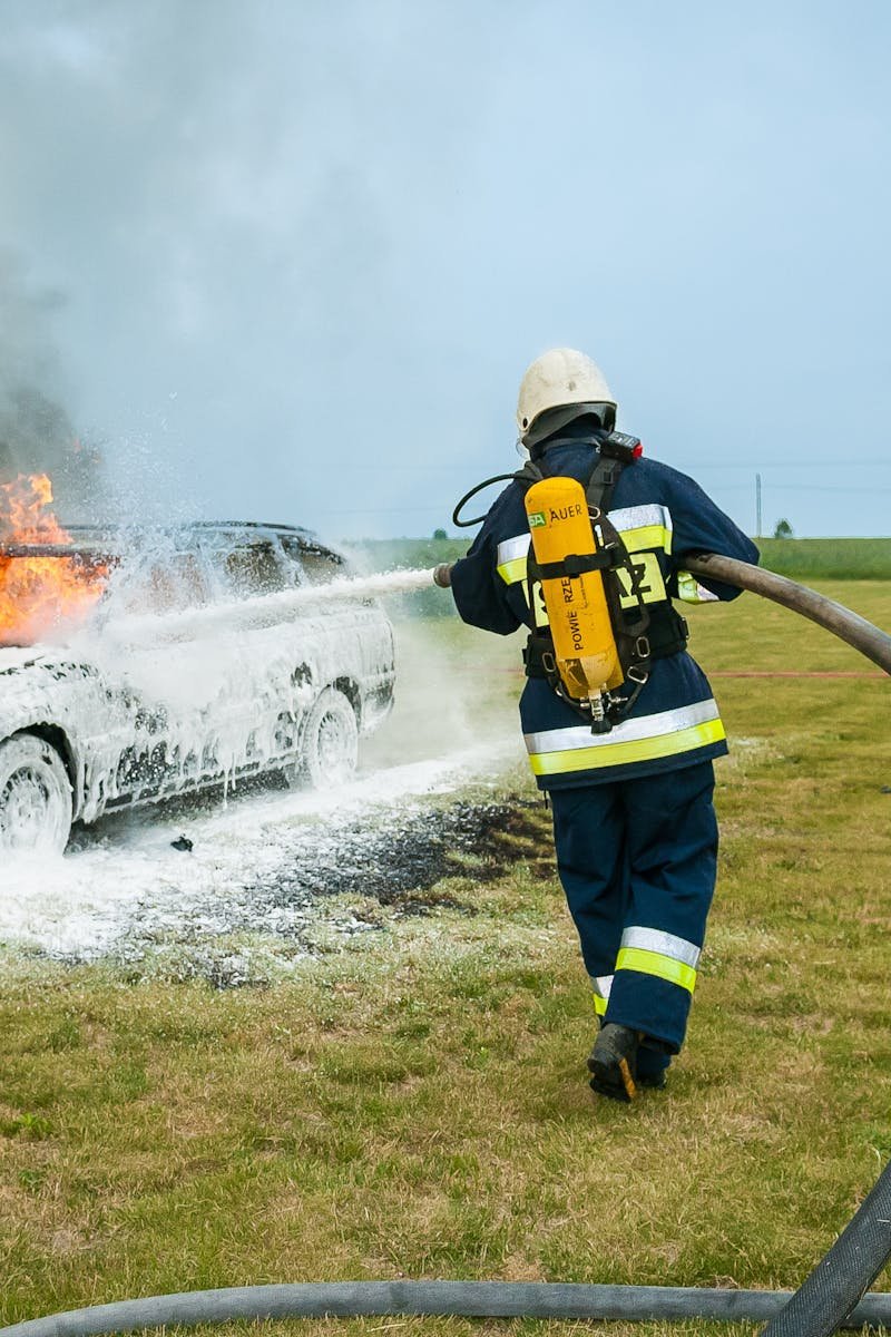 Firefighters tackle a blazing car using foam to extinguish the flames in an outdoor setting.